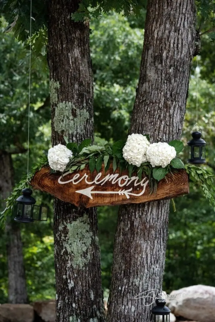 A sign with flowers on it hanging from the side of trees.